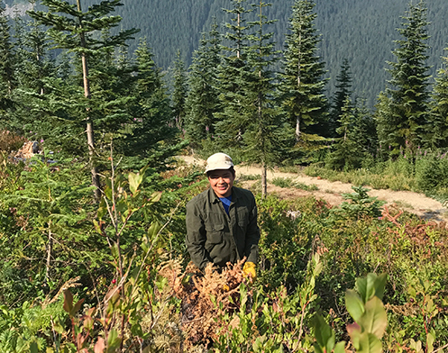 Kaiser Moses smiles as he stands in a clear cut area with growing evergreens in the mountains